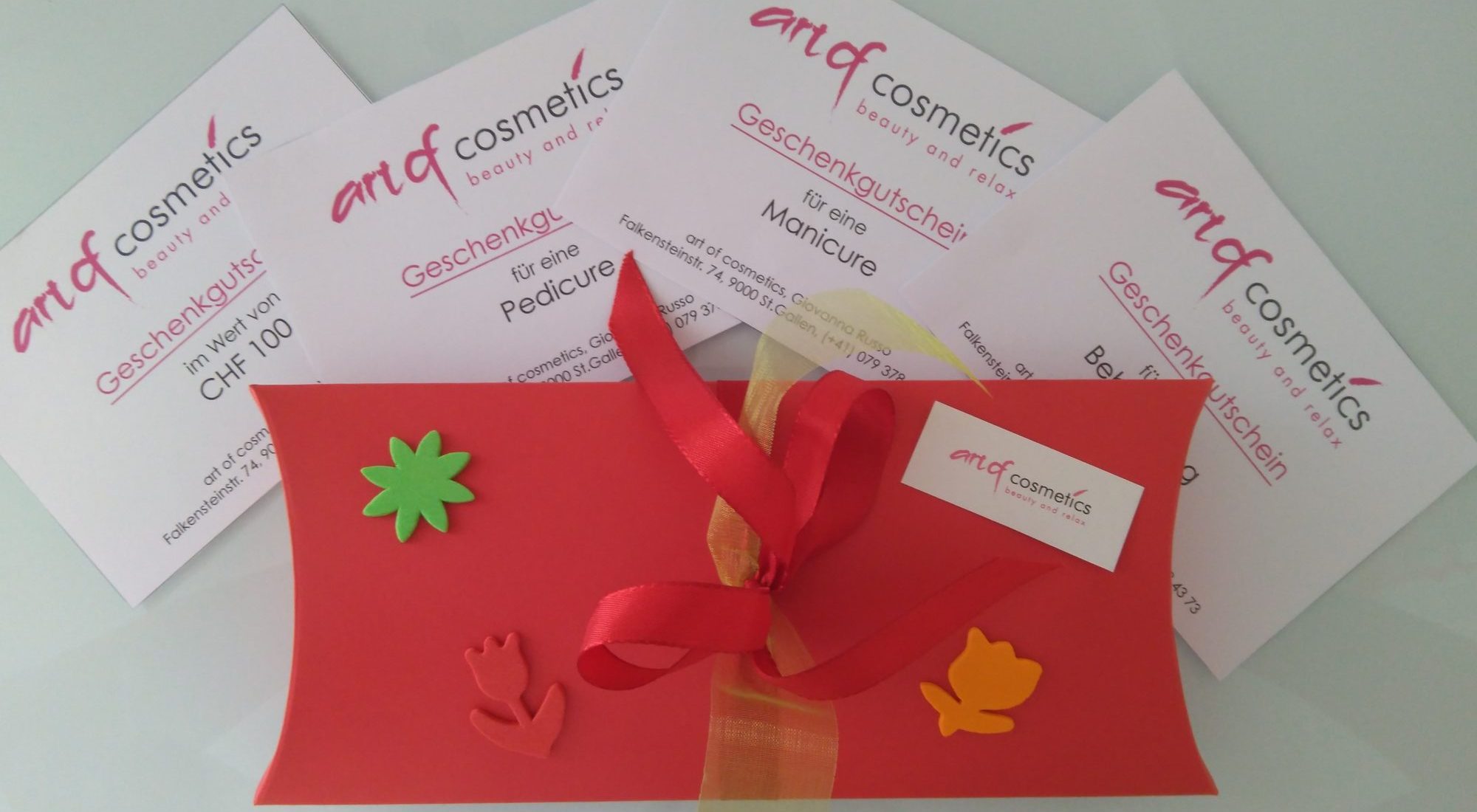 Eine rote Karte mit einer roten Schleife und einem roten Band wird gezeigt, es ist ein verpackter Geschenkgutschein von art of cosmetics in St. Gallen für eine Hochzeit oder einen besonderen Anlass. Die Karte liegt auf einem Tisch, an dem eine rote Schleife befestigt ist. Die Gestaltung der Karte und die Schleife deuten auf einen feierlichen oder festlichen Anlass hin. Im Hintergrund sind verschiedene Geschenk- und Wertgutscheine von art of cosmetics in St. Gallen zu sehen.